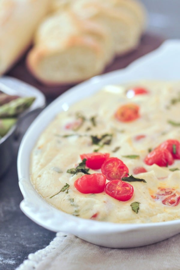 close up of melty cheese pizza dip garnished with juicy red cherry tomatoes and chopped fresh basil, in a white serving dish on a dark marbled table. sliced baguette blurred in background.