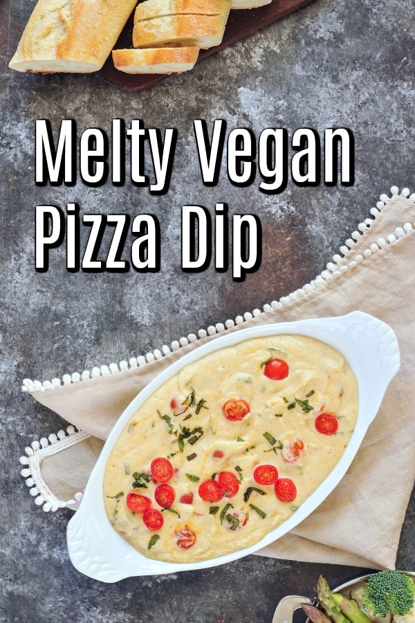 overhead view of a melty cheese pizza dip garnished with juicy red cherry tomatoes and chopped fresh basil, in a white serving dish on a dark marbled table. a silver serving dish of broccoli, cauliflower, and asparagus and a baguette serve alongside.