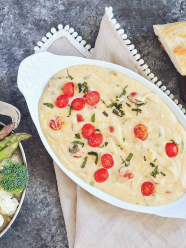 overhead view of a melty cheese pizza dip garnished with juicy red cherry tomatoes and chopped fresh basil, in a white serving dish on a dark marbled table. a silver serving dish of broccoli, cauliflower, and asparagus and a baguette serve alongside.