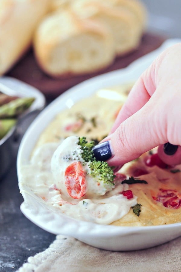 a hand dipping a fresh broccoli tree into melty cheese pizza dip garnished with juicy red cherry tomatoes and chopped fresh basil, in a white serving dish on a dark marbled table. sliced baguette blurred in background.