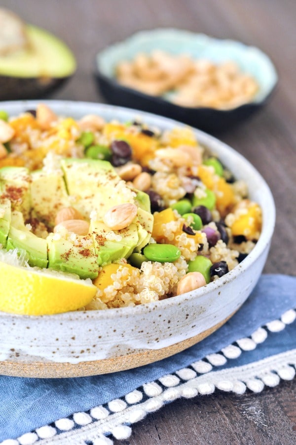 mango edamame quinoa salad with black beans, avocado, and a lemon wedge served in a shallow off white bowl. bowl is sitting on a blue cloth napkin on a wood surface.