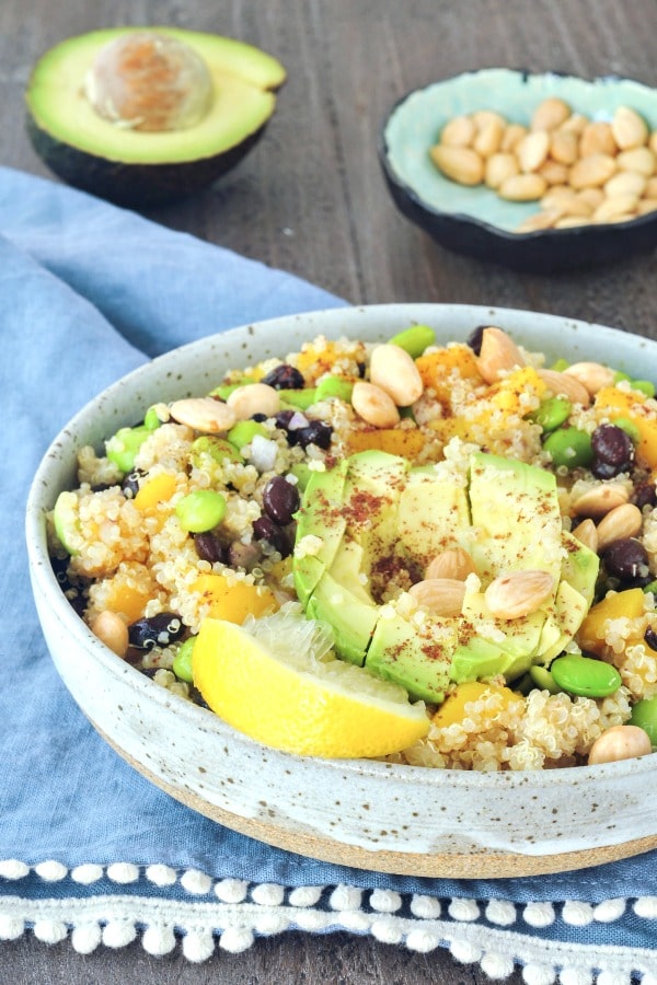 Mango Black Bean Quinoa Salad is the best potluck recipe. in a serving bowl with half a cubed avocado and a squeezed slice of lemon on top. the other half of avocado and a small bowl of almonds in background.