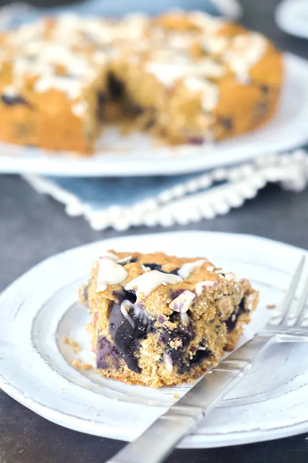 one slice of Blueberry Breakfast Cake on a plate with a fork, full cake on platter in background