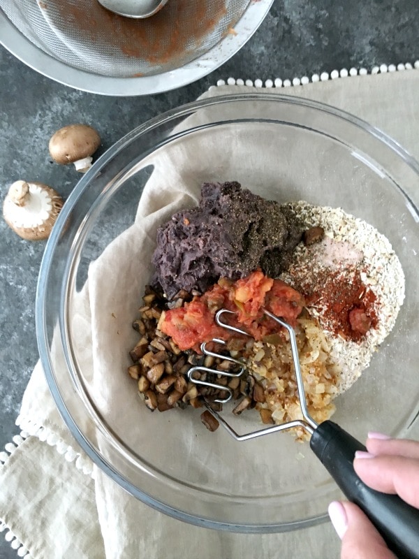 How To Make A Veggie Burger That Doesn't Fall Apart: glass bowl with veggie burger ingredients - black beans, mushrooms, oats, salsa, spices, being mixed by a hand holding a masher