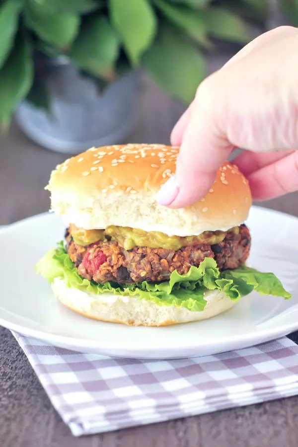 A hand is picking up a southwest black bean burger dressed with lettuce, cheese sauce, green chiles and a sesame seed bun.