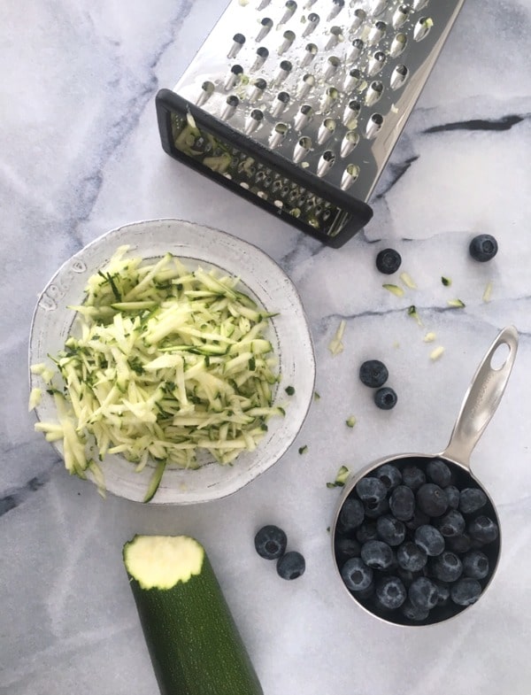 Vegan Zucchini Bread Recipe: a plate of grated zucchini next to a box grater, a cup of fresh blueberries