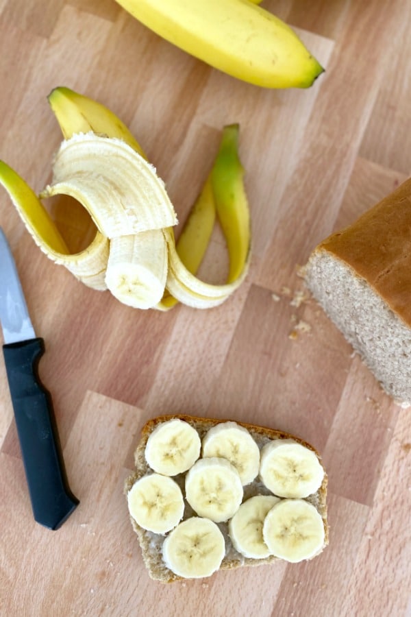 one slice of Gluten Free Peanut Butter Bread with slices of banana on a cutting board, more banana and loaf of bread on the side