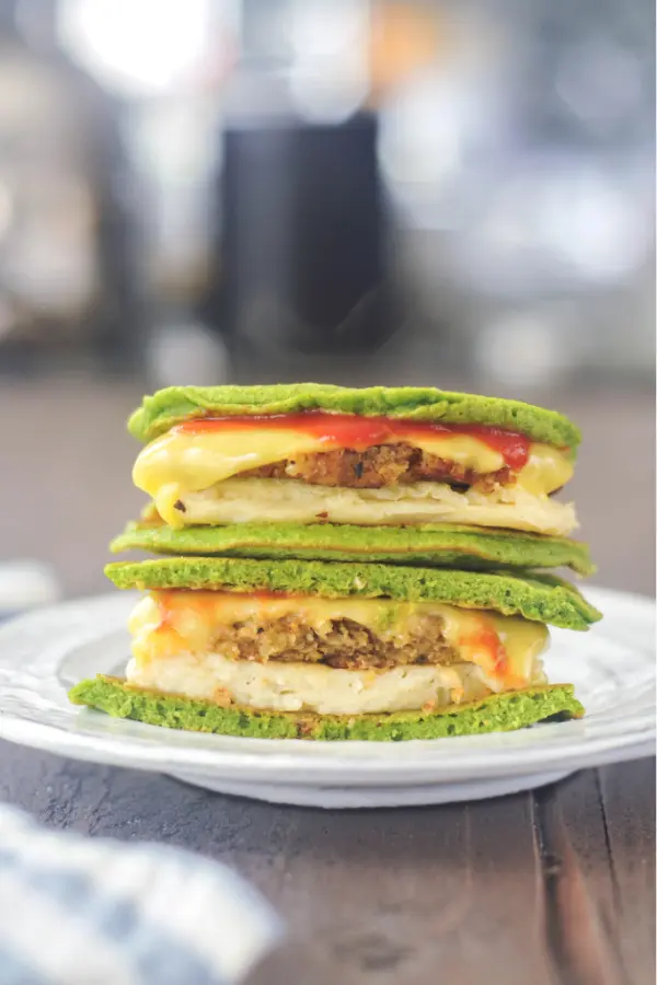 a breakfast sandwich using green vegan spinach pancakes as bread. sliced in half and stacked on a plate to show filling: vegan egg, sausage, melted cheese, ketchup between bright green pancakes.