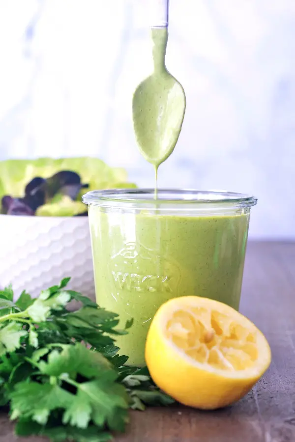 a jar of Green Goddess dressing with a spoon coming out of it, a squeezed lemon and a bunch of herbs on the side.