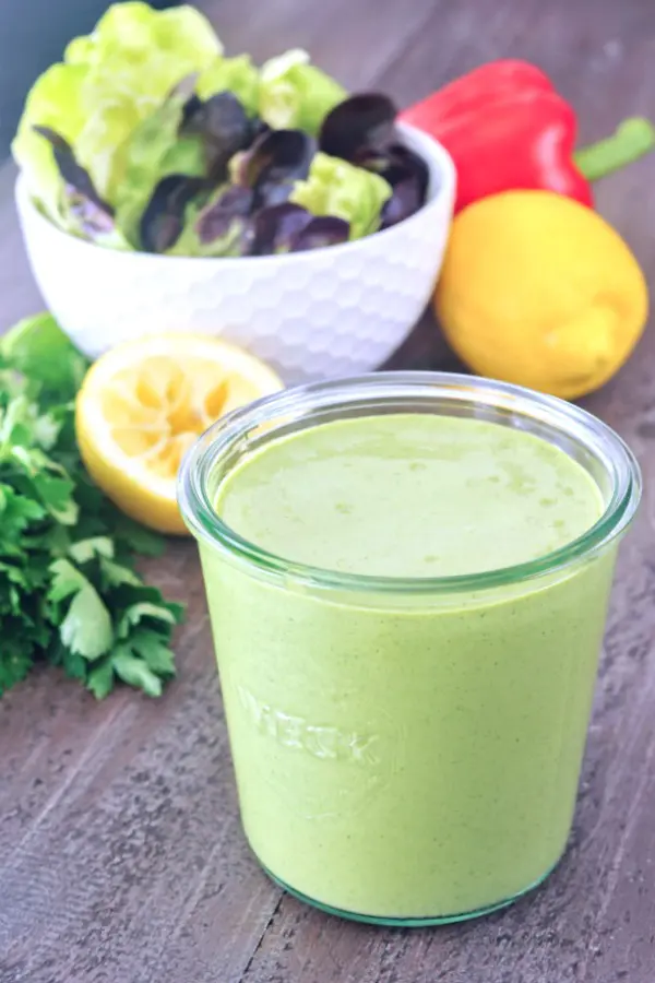 a jar of Green Goddess dressing in front of a salad bowl of greens. a squeezed lemon, a red bell pepper, and a bunch of fresh herbs in background.