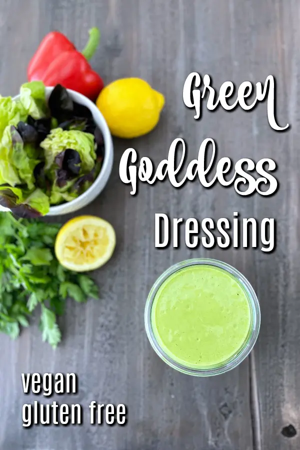 overhead photo of a jar of Green Goddess dressing and a salad bowl of greens. a squeezed lemon, a red bell pepper, and a bunch of fresh herbs in background.