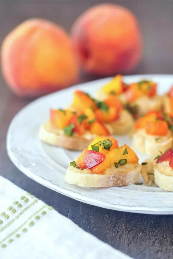 rustic plate with sweet cream peach crostini topped with chopped basil leaves, two fresh peaches in background