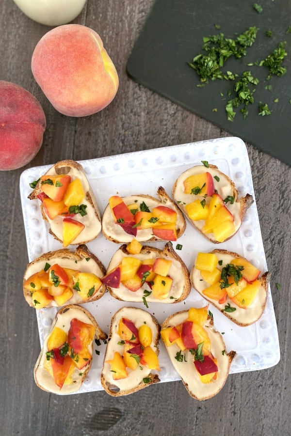 overhead view of roasted peach crostini arranged on a square white dish: toasted baguette slices with sweet cream, roasted peaches, and fresh chopped basil