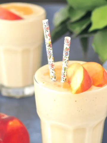 two glasses of peach smoothie, garnished with fresh peach slices and cinnamon. one glass has floral decorated paper straws, and a fresh peach sliced in half next to the glass, green plant in background