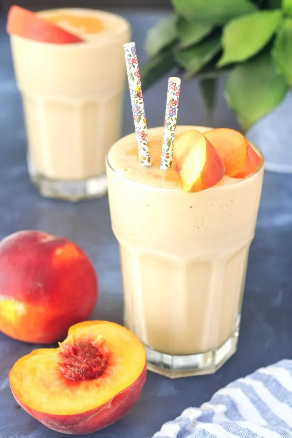 two glasses of peach smoothie, garnished with fresh peach slices and cinnamon. one glass has floral decorated paper straws, and a fresh peach sliced in half next to the glass, green plant in background