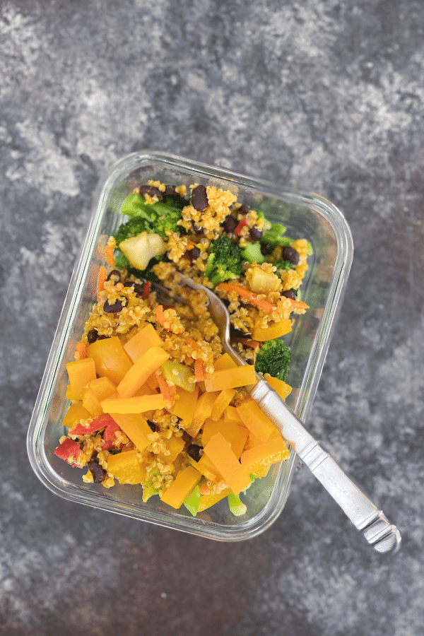 ginger carrot flavored quinoa with black beans and veggies in a single serve, glass meal prep container. on dark grey background, sliver matte finish fork in container.