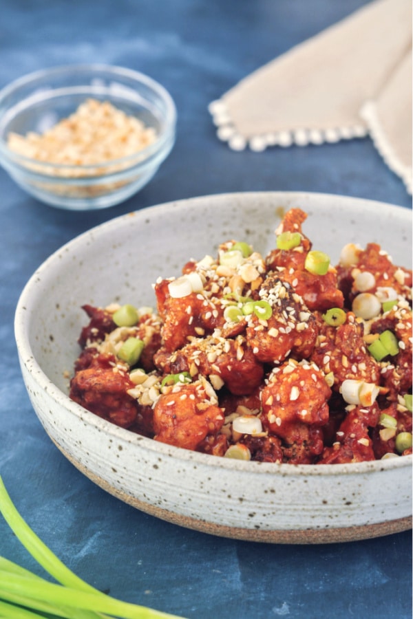 shallow bowl of dark orange spicy air fried cauliflower florets, garnished with green onion and sesame seeds.