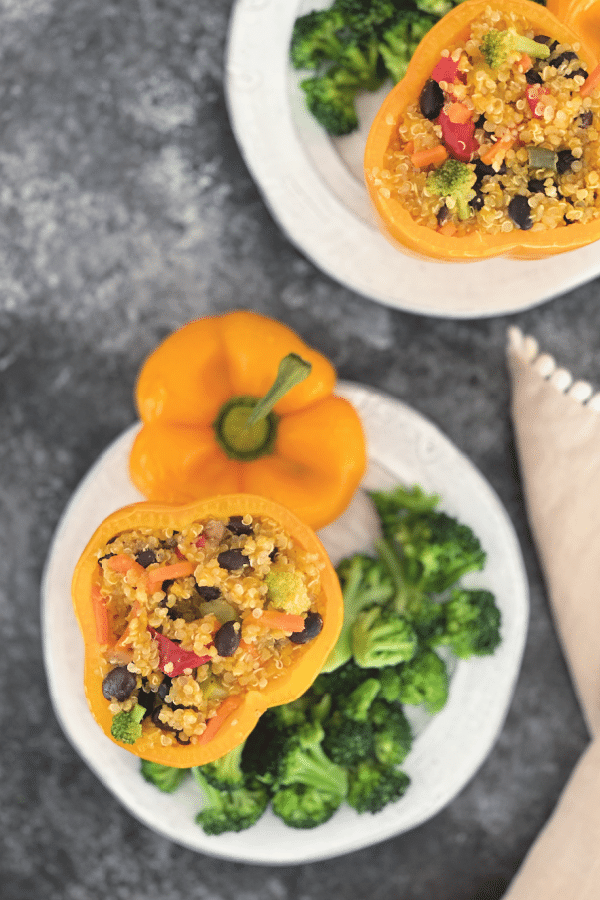 overhead view of two orange bell peppers on separate plates, stuffed with ginger carrot quinoa, black beans, and broccoli, on a white plate with more broccoli on the side 