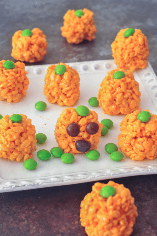 a large platter of vegan rice crispy treats colored orange and rolled into pumpkin shapes for Halloween. green candy on top for pumpkin stem. more green candy scattered on the platter, for pumpkin patch greenery.