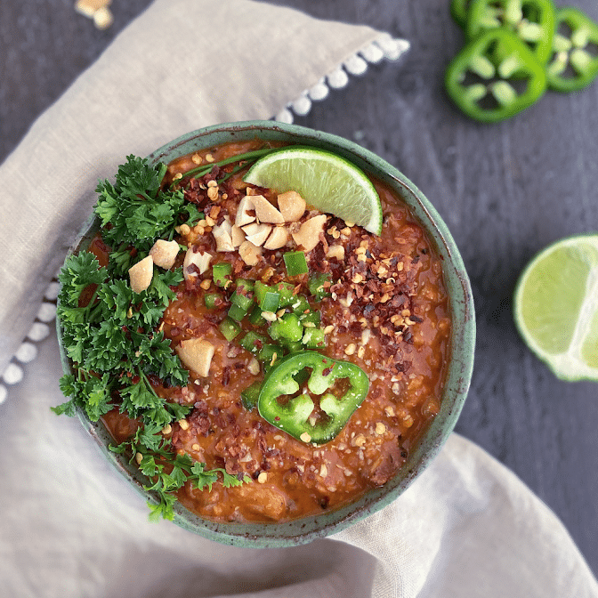 overhead view of bright orange spicy vegan peanut soup in a rustic bowl, topped with parsley, sliced jalapeno, chopped peanuts.