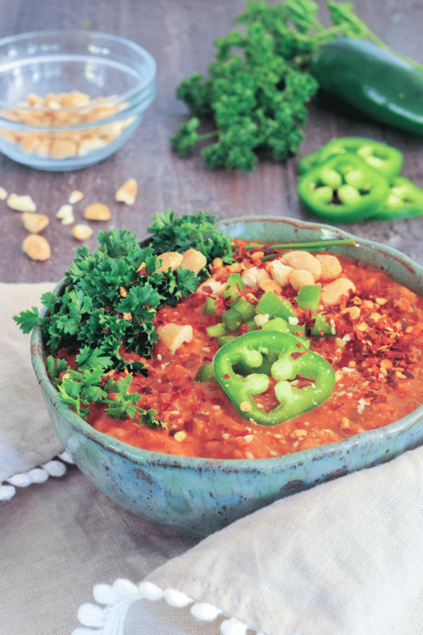 bright orange spicy vegan peanut soup in a rustic grey pottery bowl, topped with parsley, sliced jalapeno, chopped peanuts. bowl of peanuts, whole jalapeno pepper, parsley in the background on a wood table, beige napkin in foreground in front of soup bowl.