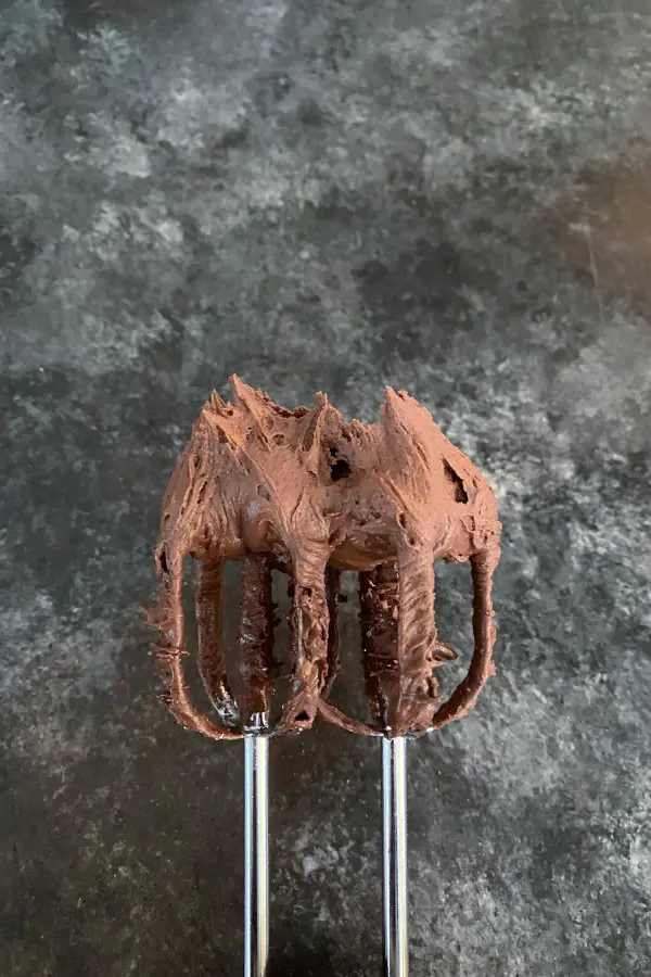 Two mixer beaters with chocolate fudge frosting on them, standing up in stiff peaks.