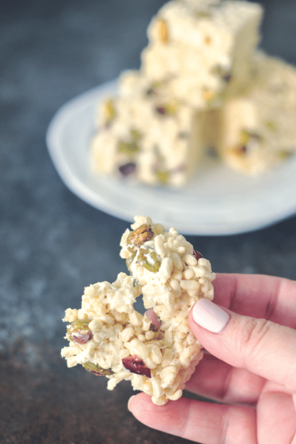 Cranberry Pistachio Rice Crispies pulled apart in a hand, a plate of rice crispies blurred in the background.