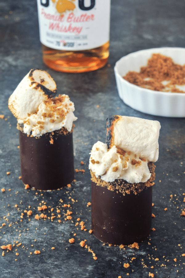 s'mores shots in edible chocolate shot glasses, topped with whipped cream, cookie crumbs, and a toasted marshmallow. whiskey bottle and shallow bowl with cookie crumbs in (dark) background.