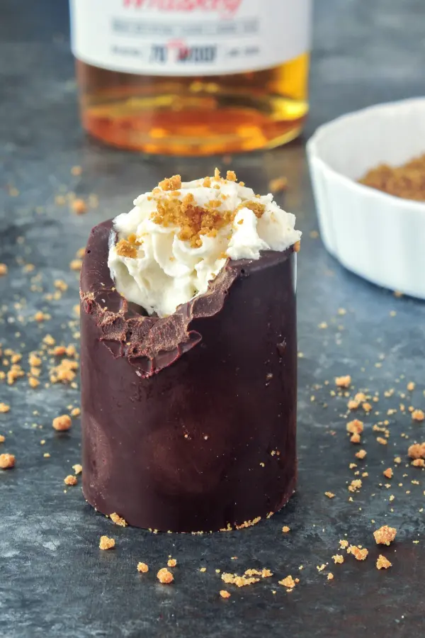 a chocolate shot glass with a bite out of it, filled with whipped cream and topped with cookie sprinkles. whiskey bottle in background, dark surface.