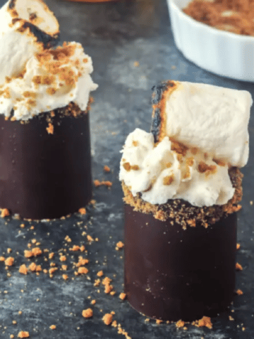 s'mores shots in edible chocolate shot glasses, topped with whipped cream, cookie crumbs, and a toasted marshmallow. whiskey bottle and shallow bowl with cookie crumbs in (dark) background.