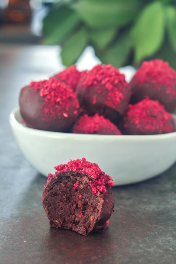 chocolate truffles with freeze dried raspberry dust in a white bowl, one out of the bowl and sliced in half to show the brownie filling.