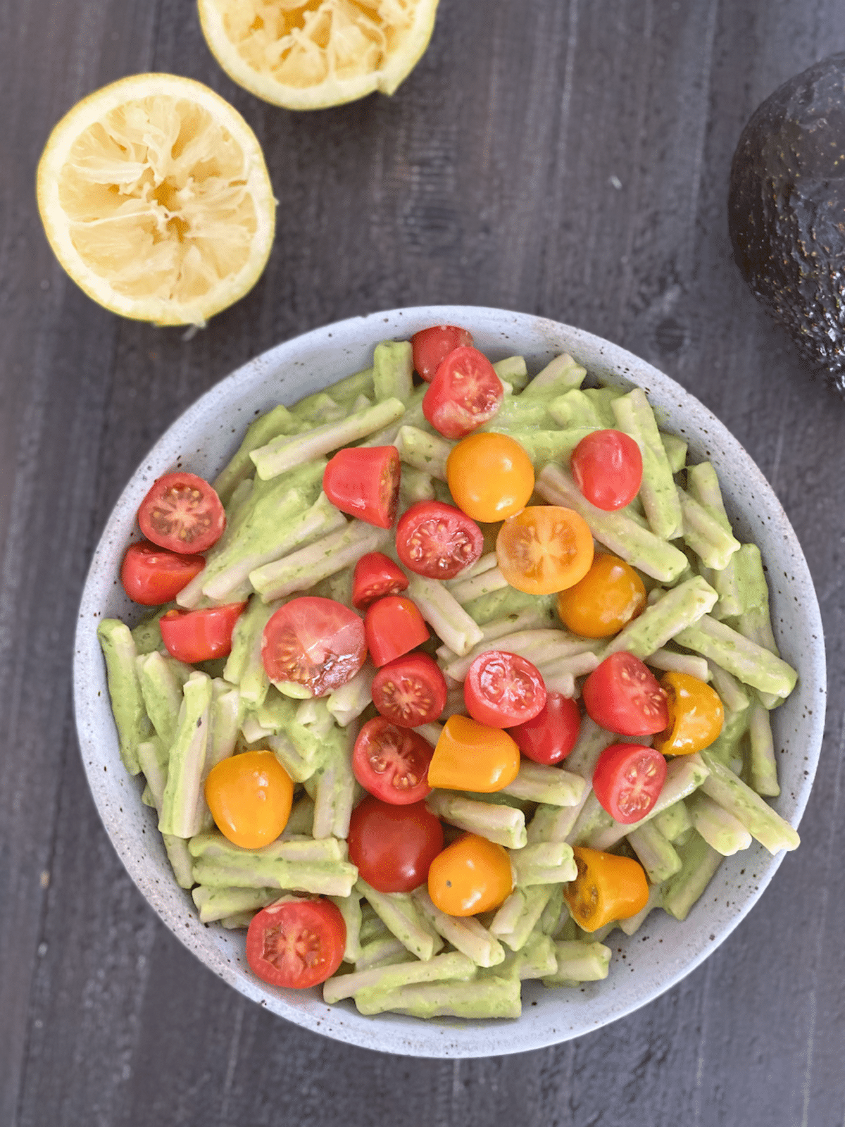 an overhead view of a bowl of pasta with light green avocado sauce and sliced tomatoes