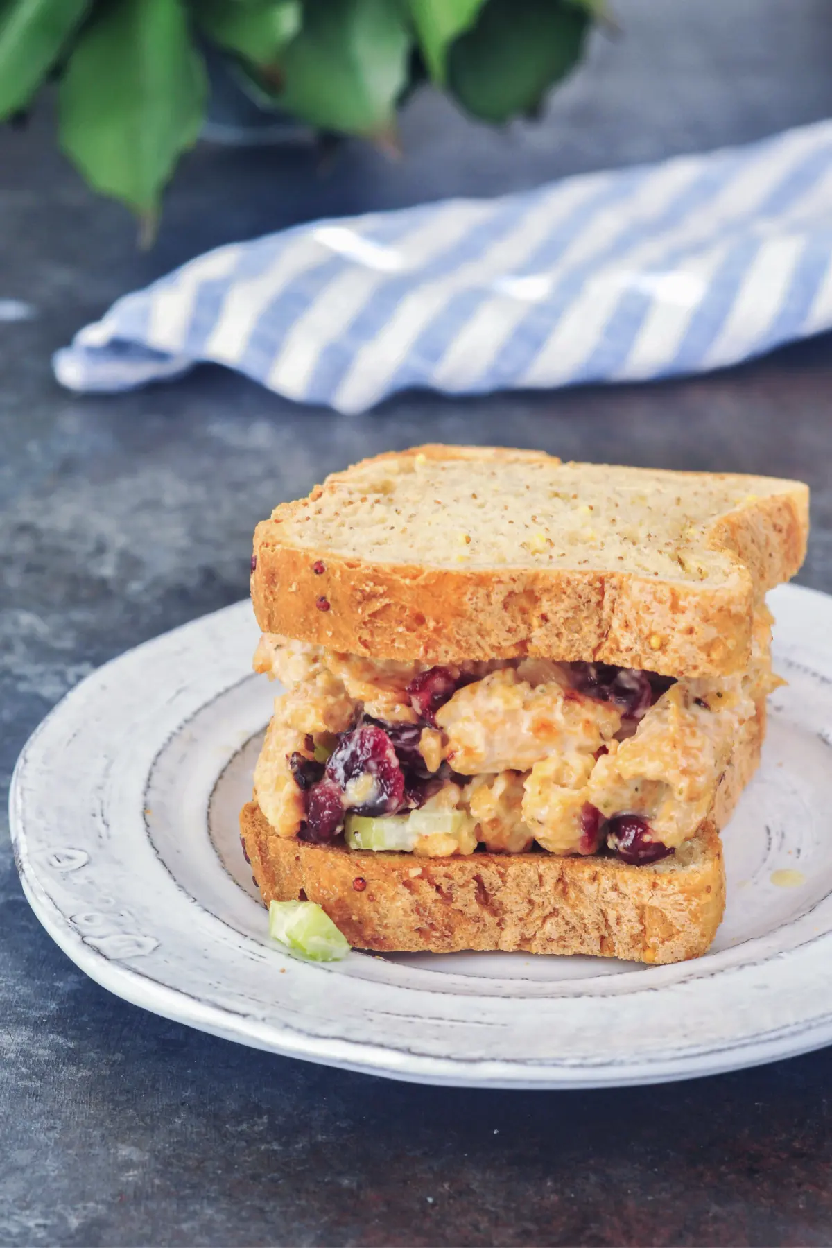 a vegan chicken salad sandwich on a rustic white dish, blue and white striped napkin in background. vegan chicken salad is made from soy curls, and has chopped celery and dried cranberries. 