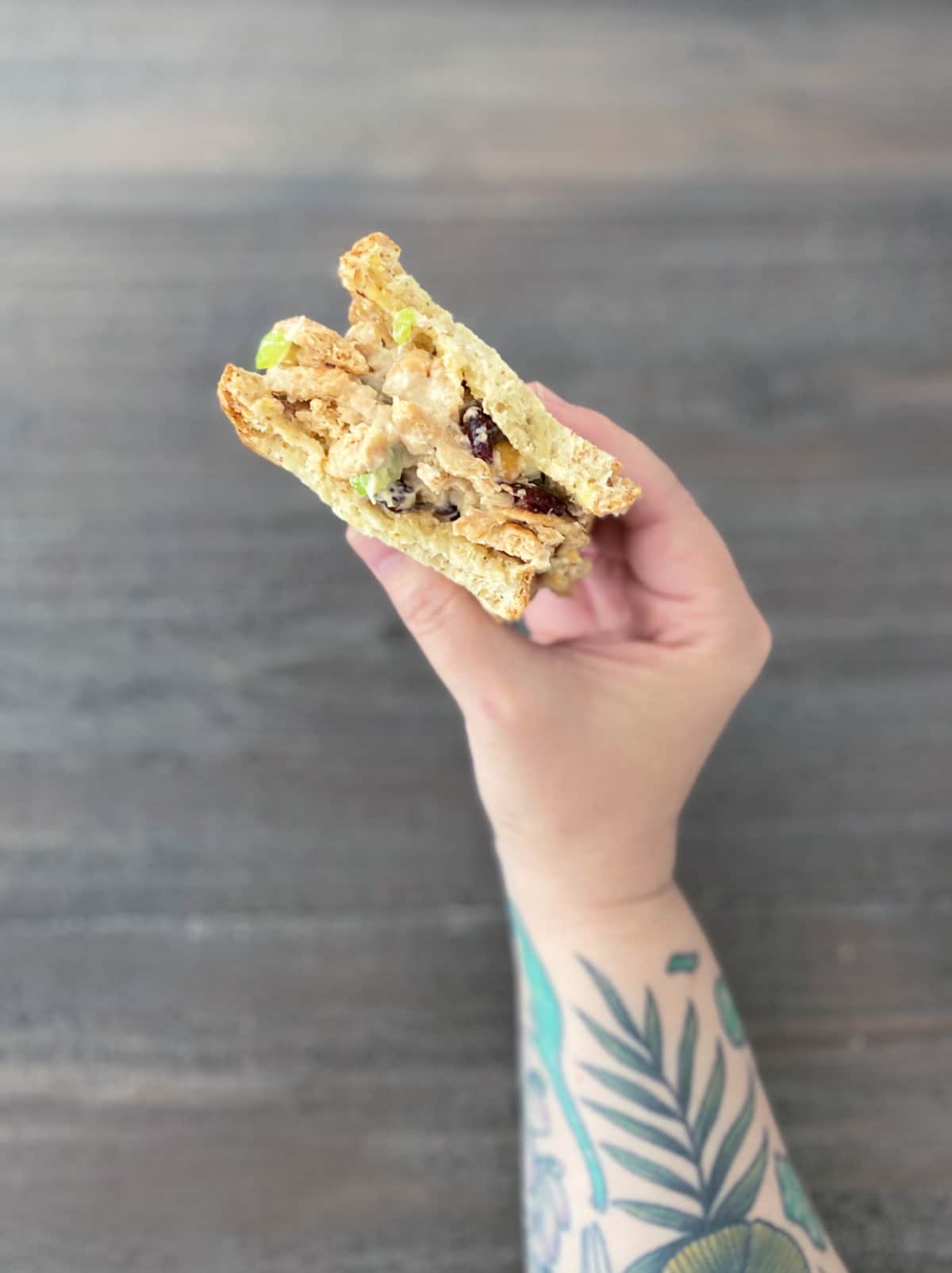 a hand (with leafy green tattoos on arm) holds a vegan chicken salad sandwich over a wood table. vegan chicken salad is made from soy curls, and has chopped celery and dried cranberries. 