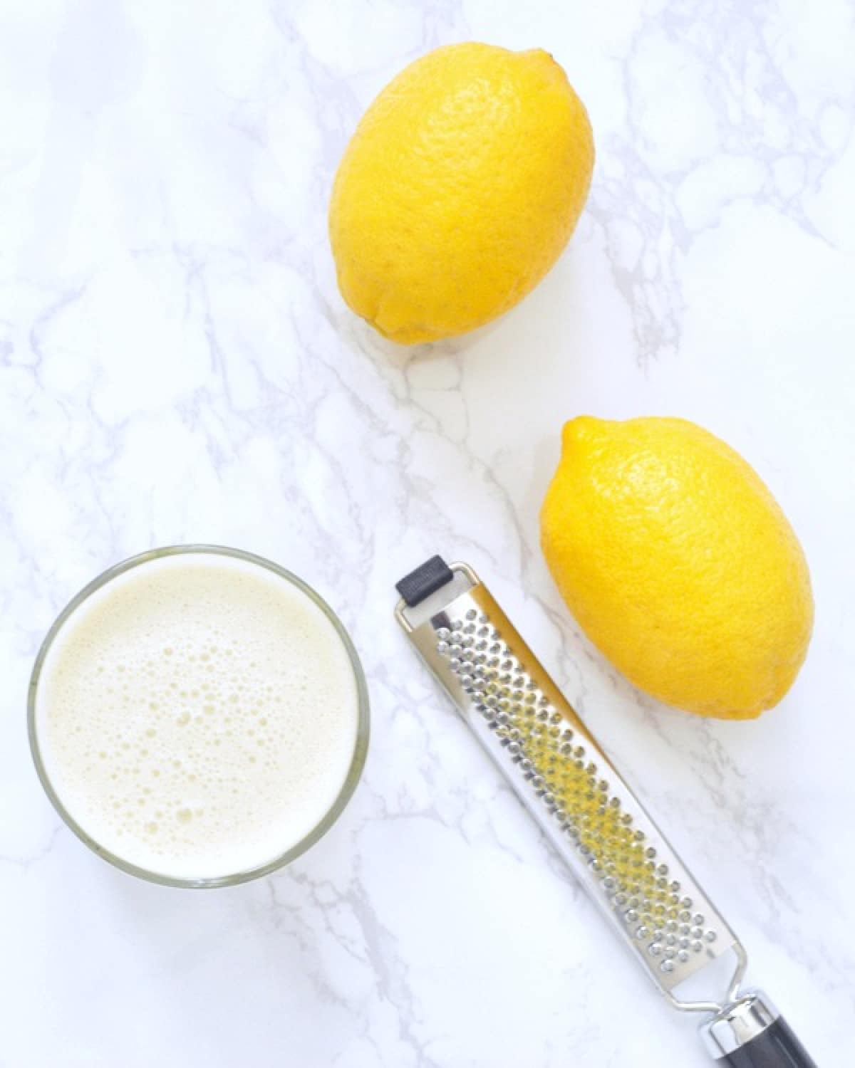 an overhead view of a lemon smoothie in a glass on a white marble counter. two fresh lemons and a zester sit next to the smoothie.
