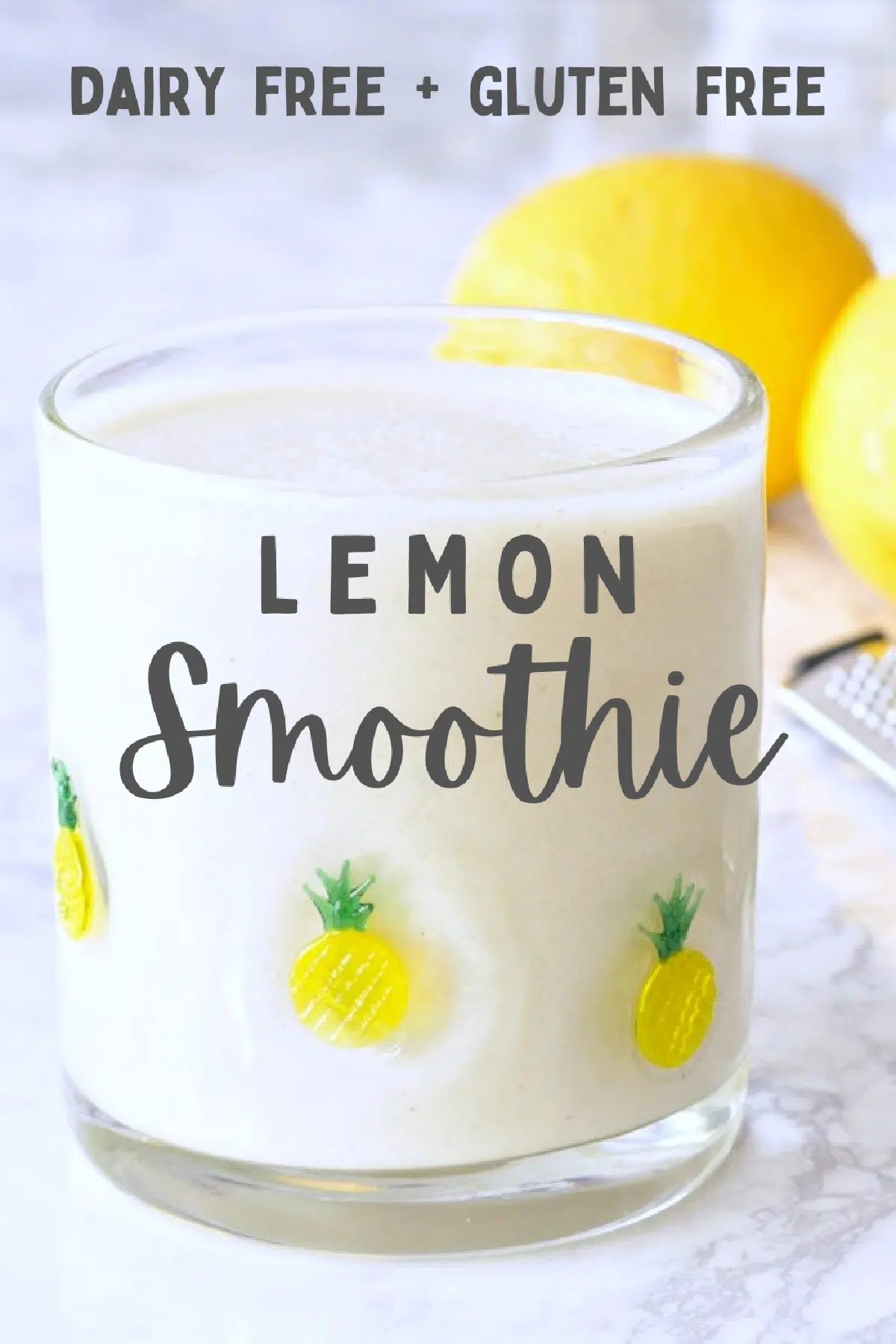 a short clear glass filled with a bright white lemon smoothie. two fresh lemons and a zester sit behind the smoothie.