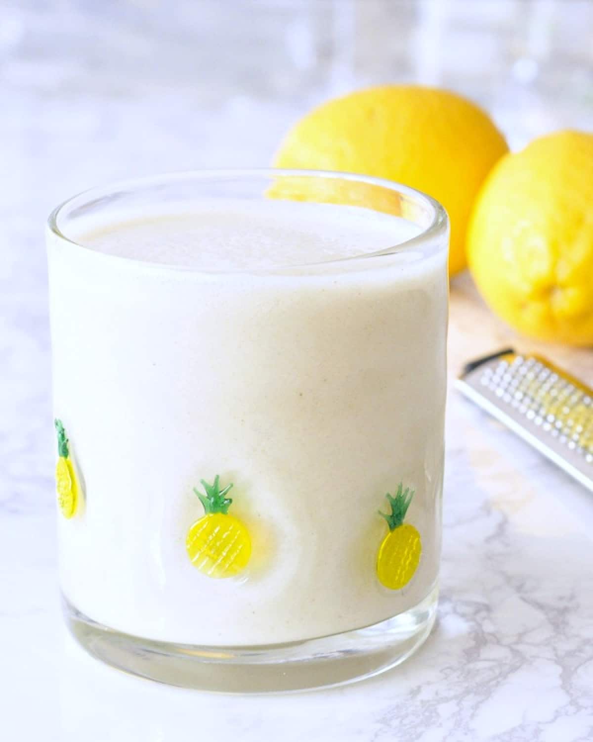 a short clear glass filled with a lemon smoothie that is white in color. two fresh lemons and a zester sit behind the smoothie.