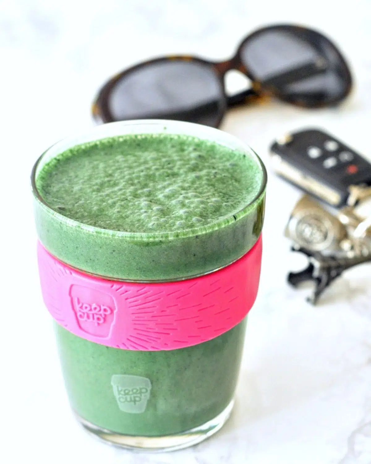 a green lemon spinach smoothie in a glass travel cup on a white marble countertop; car keys and sunglasses in background.