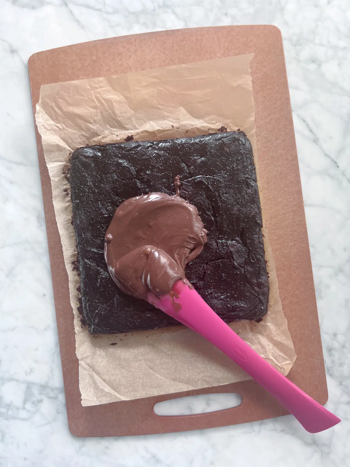 black bean brownies before slicing, on parchment and cutting board. a red spatula sits in chocolate icing that hasn't been spread over the brownies yet.