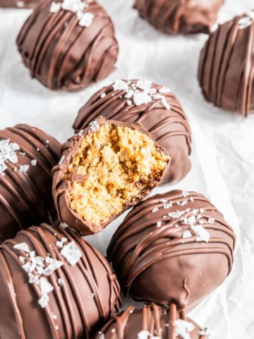 chocolate peanut butter balls with flake sea salt sitting on a piece of white parchment, one ball sliced open to show crispy peanut butter filling inside the chocolate coating