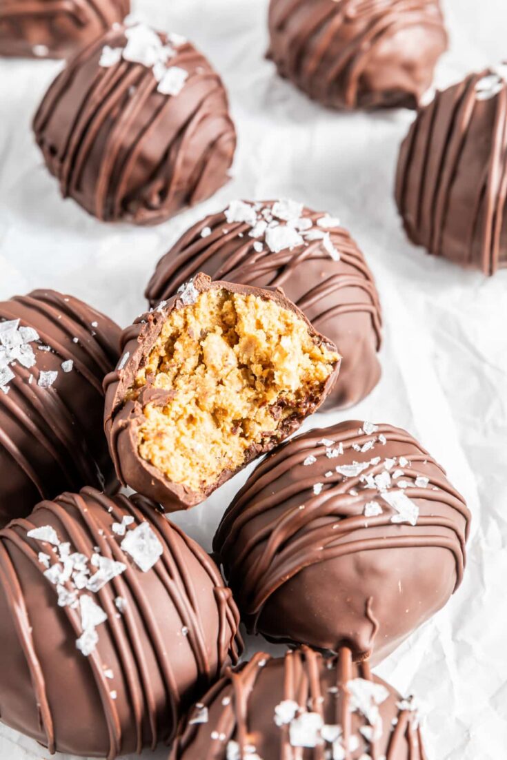 chocolate peanut butter balls with flake sea salt sitting on a piece of white parchment, one ball sliced open to show crispy peanut butter filling inside the chocolate coating