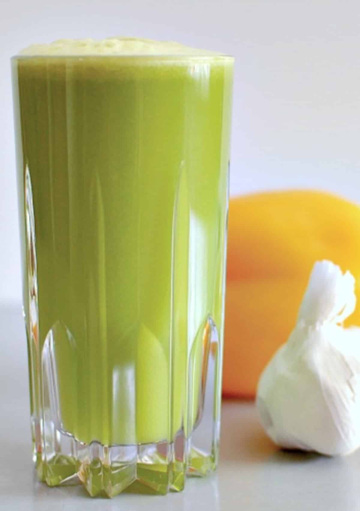 side view of bright green garlic apple juice in a tall cut crystal glass. juice has light green foam on top, and there is a yellow bell pepper and a white bulb of garlic sitting beside the glass of juice.