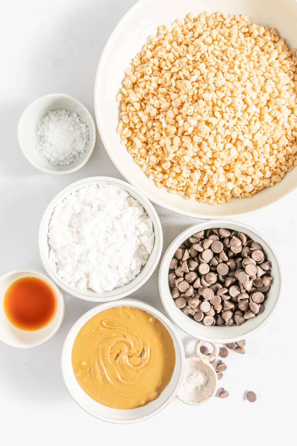 the ingredients for butterfinger balls measured out in separate bowls, ready to make: crispy rice cereal, chocolate chips, powdered sugar, peanut butter, vanilla extract, flaked sea salt