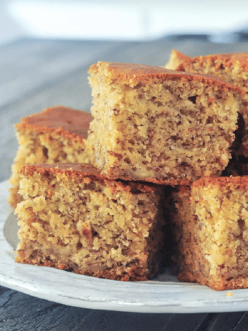 squares of banana snack cake stacked on a rustic dish, on a wooden table against a blurred kitchen background.