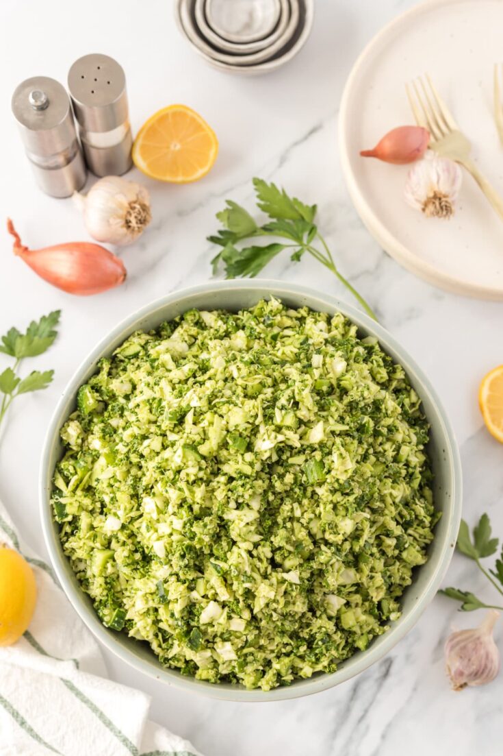 overhead view of green goddess chopped salad in a bowl with some of the ingredients in the background: a shallot, herbs, lemon slices