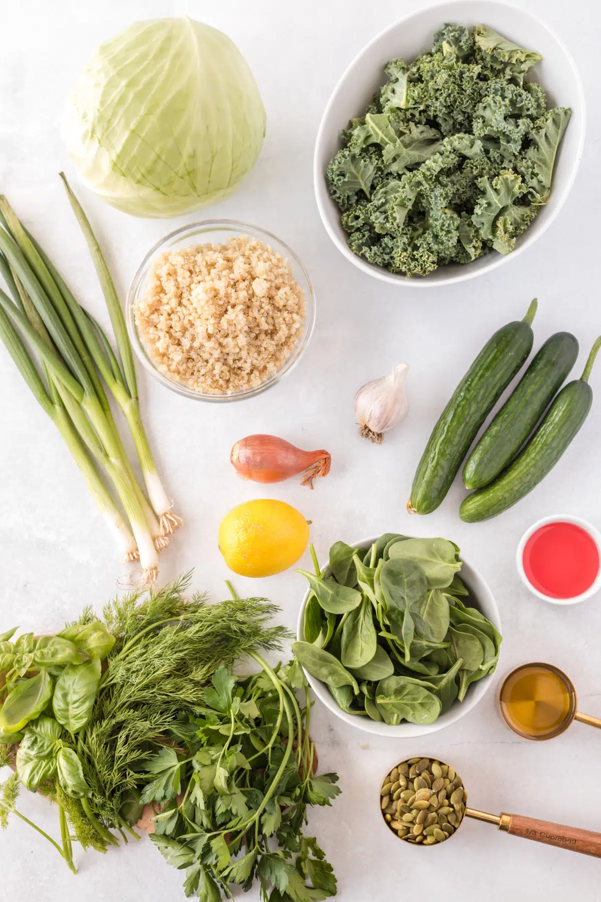 overhead view of all green goddess salad ingredients, before prepping: whole cucumbers, green onions, kale leaves, head of cabbage, whole lemon, nuts, olive oil, all on a white background