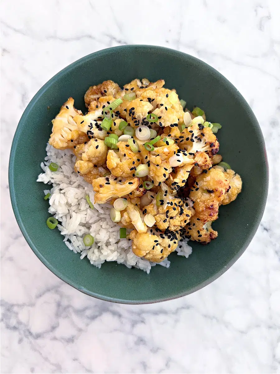 overhead view of orange cauliflower florets over rice in a shallow dark green bowl, garnished with sliced green onion and black sesame seeds