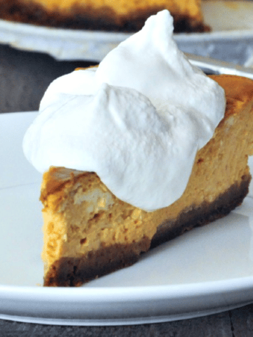 a slice of pumpkin cheesecake on a white plate with bright white whipped cream being spooned on top; the remaining whole cheesecake in background.