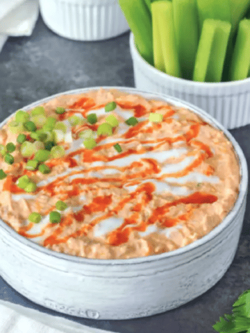 Vegan buffalo chicken dip in a large straight sided, rustic grey serving bowl, garnished with hot sauce and sour cream drizzle and sliced green onion. set on a dark grey marble tabletop, with smaller bowls of celery slices for dipping.