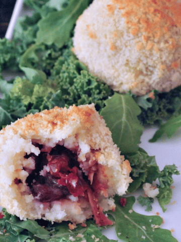 an inside view of crispy vegan stuffed potato balls - sitting on curly kale on a white plate, this panko crusted mashed potato ball is cut in half to show the jackfruit filling